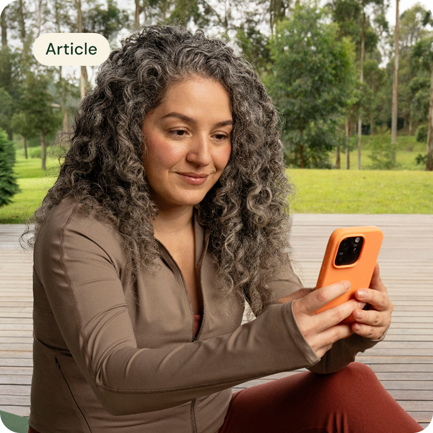 A woman is looking down at her phone outdoors, trees, grass and plants behind her.