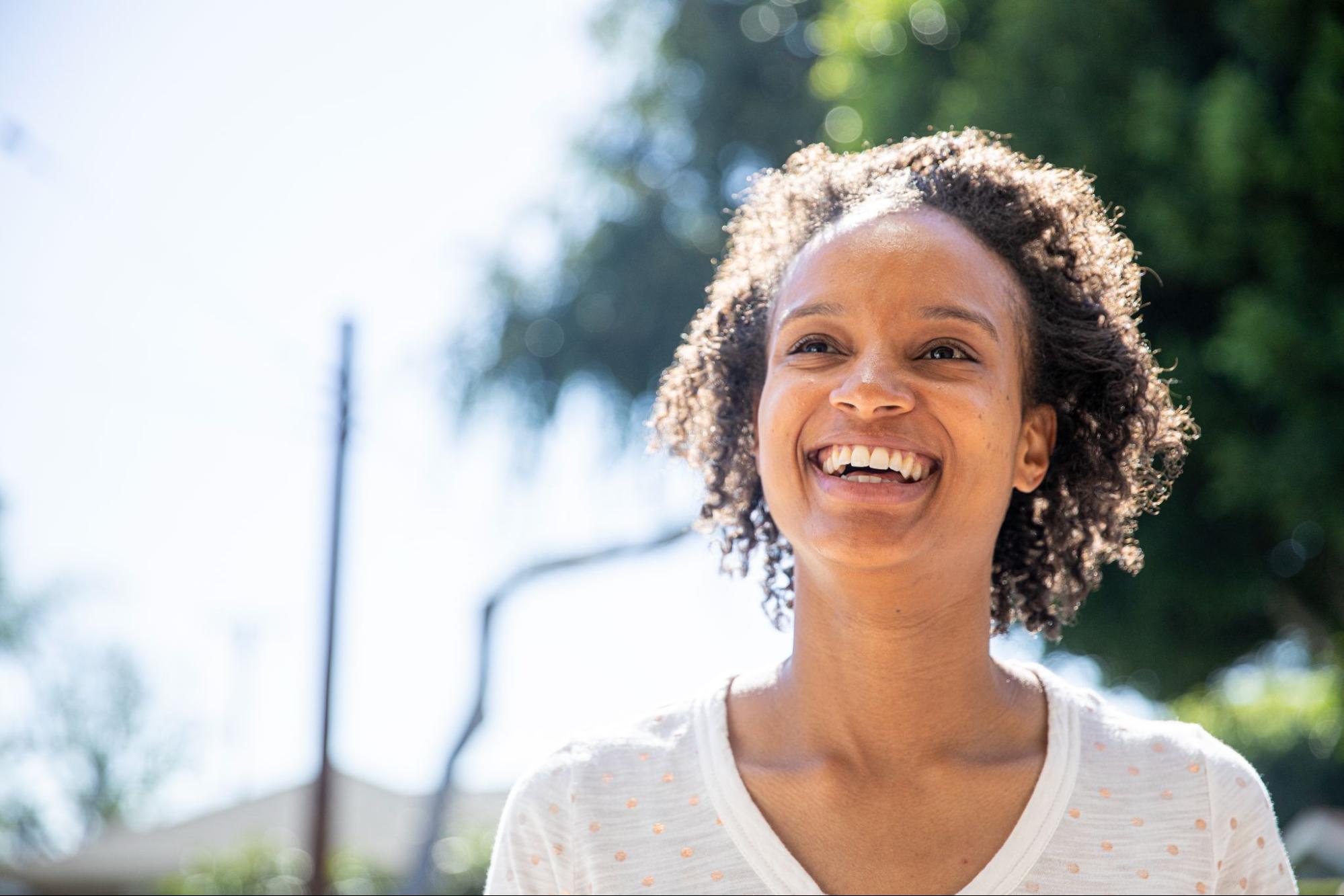 Person with natural curly hair laughing joyfully because she knows her worth, outdoors on sunny day, wearing white polka dot top