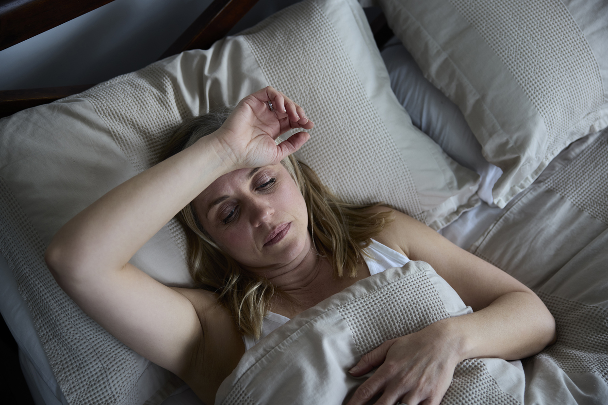Person with menopause lying in bed with hand on forehead trying to sleep, surrounded by white textured bedding