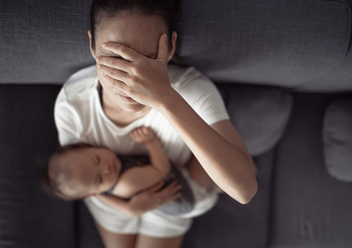 Mom in white clothing sitting in dark room covering face with hand due to postpartum headaches. 