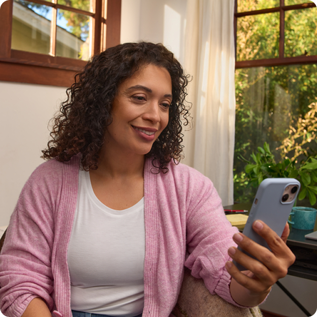 A woman sits by a sunlit window and smiles as she looks at her smartphone.
