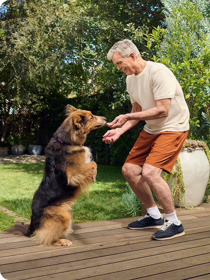 An older man bends down to pet his dog in his sunlit backyard.
