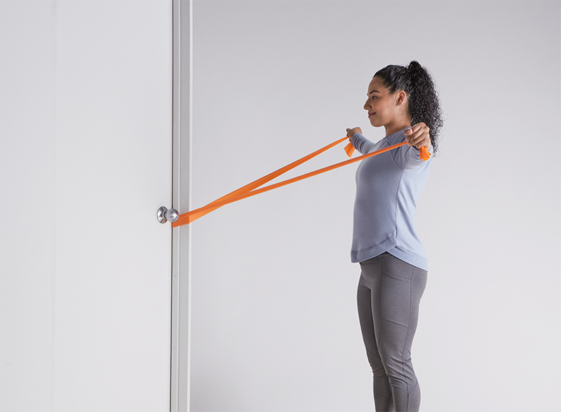 Woman in workout attire performing deltoid exercises with orange resistance band attached to door handle in bright white room