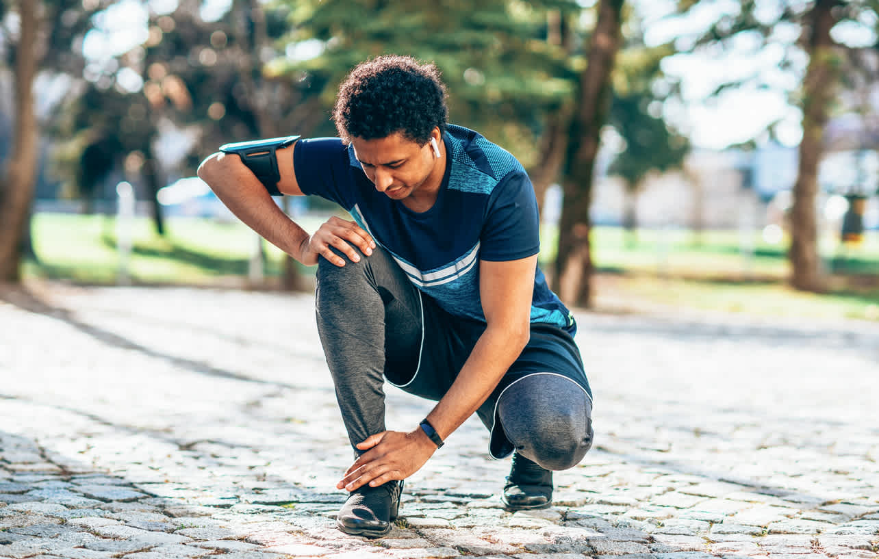 Man-kneeling-down-touching-his-ankle-mid-run-outdoors