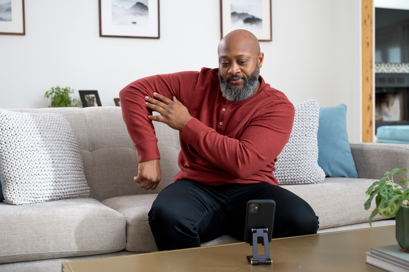 Photo of a man sitting on a couch, gesturing toward his elbow, and gazing into his phone which is set in a phone holder in front of him