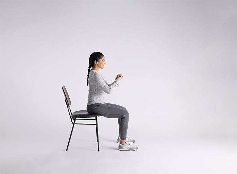 Woman in gray athletic wear performing seated bowel incontinence exercises on a black chair against white background