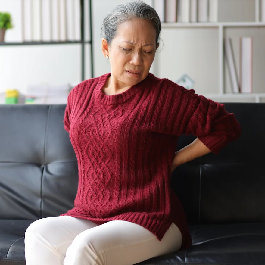 Senior woman in red sweater sitting on black couch grimacing while holding her lower back due to thoracic spondylosis.