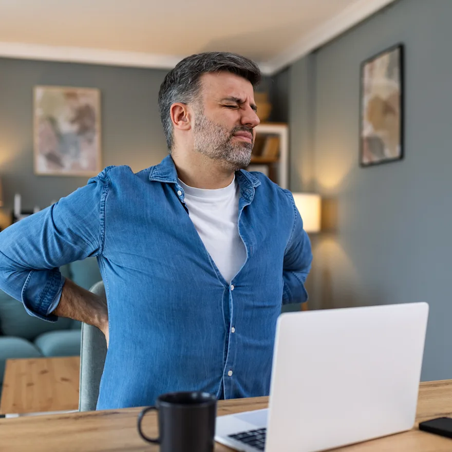 Person in blue denim shirt grimacing while holding lower back in pain while working at desk with laptop in home office setting