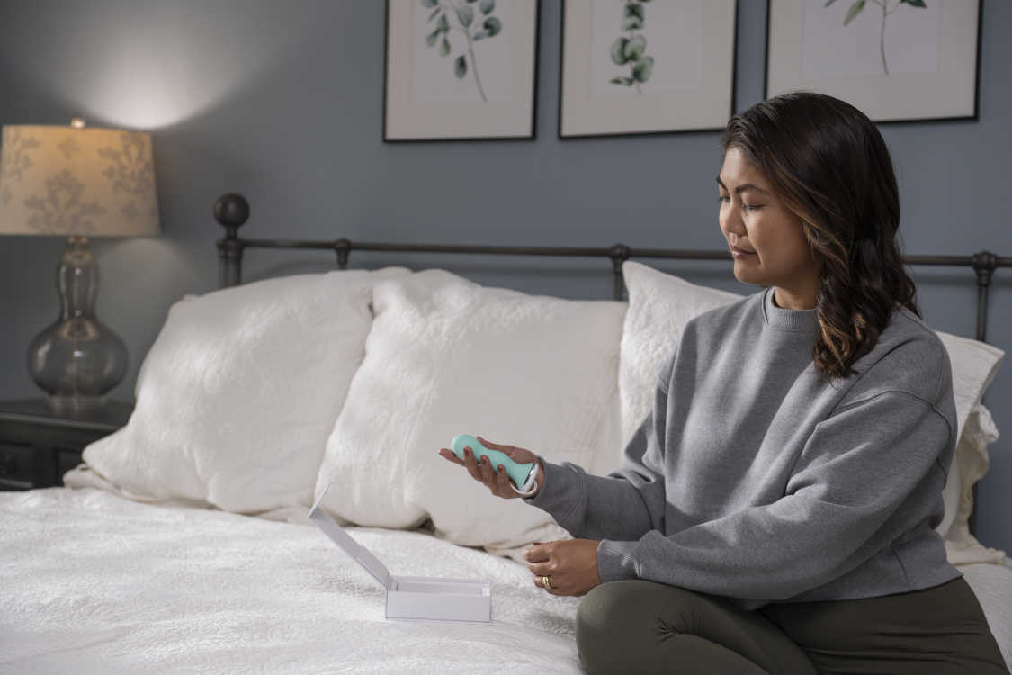 Image of a woman sitting on the edge of her bed, looking at a pelvic trainer in her hand