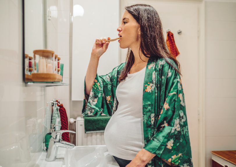 Pregnant woman in floral green robe brushing teeth in bathroom, wearing white tank top, standing at sink with mirror visible