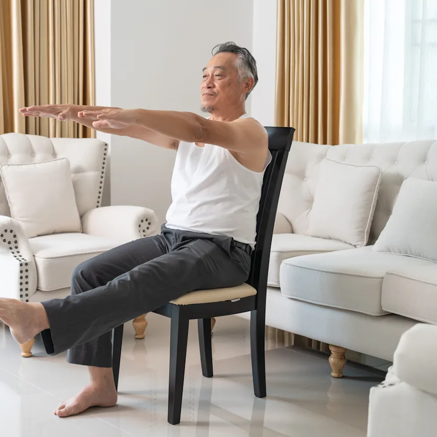 Senior man performing iliotibial band syndrome exercises, extending arms and leg while sitting on a black chair in bright living room