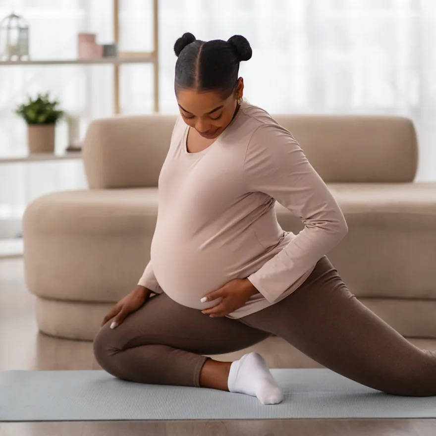 Pregnant woman in pink top and brown leggings stretching on yoga mat in bright living room with beige couch and exercise ball