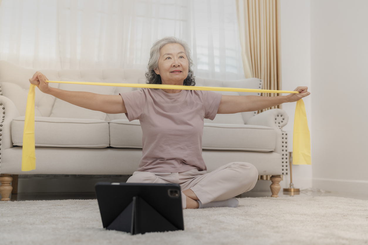 Senior woman exercising at home with yellow resistance band while seated on floor near white couch