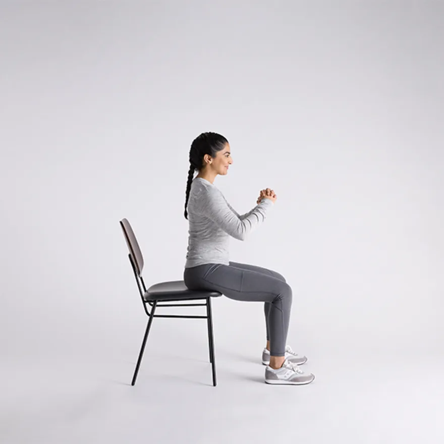 Woman in gray athletic wear demonstrating bowel incontinence exercises on a black chair against white background