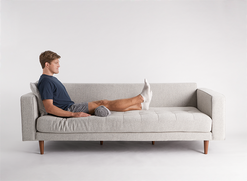 Person in navy t-shirt lying on light gray modern sofa with rolled towel under knees, practicing hip replacement exercises