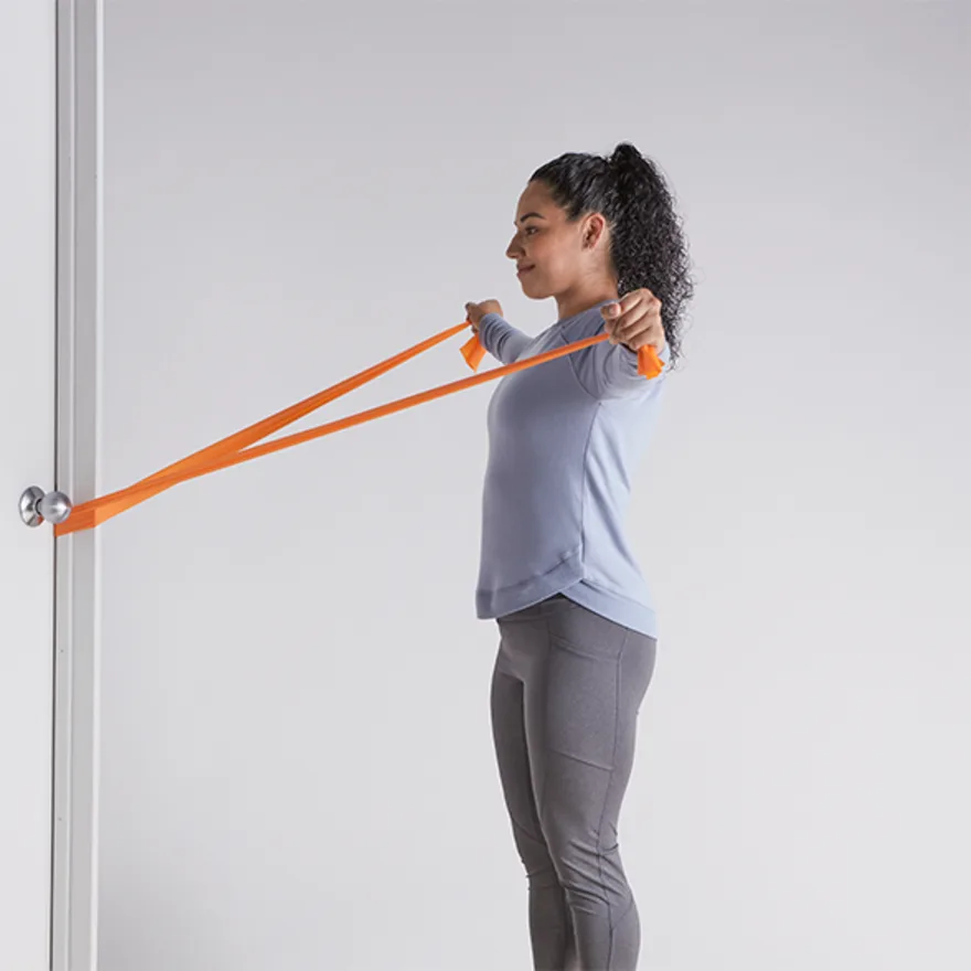 Woman in workout attire performing deltoid exercises with orange resistance band attached to door handle against white wall