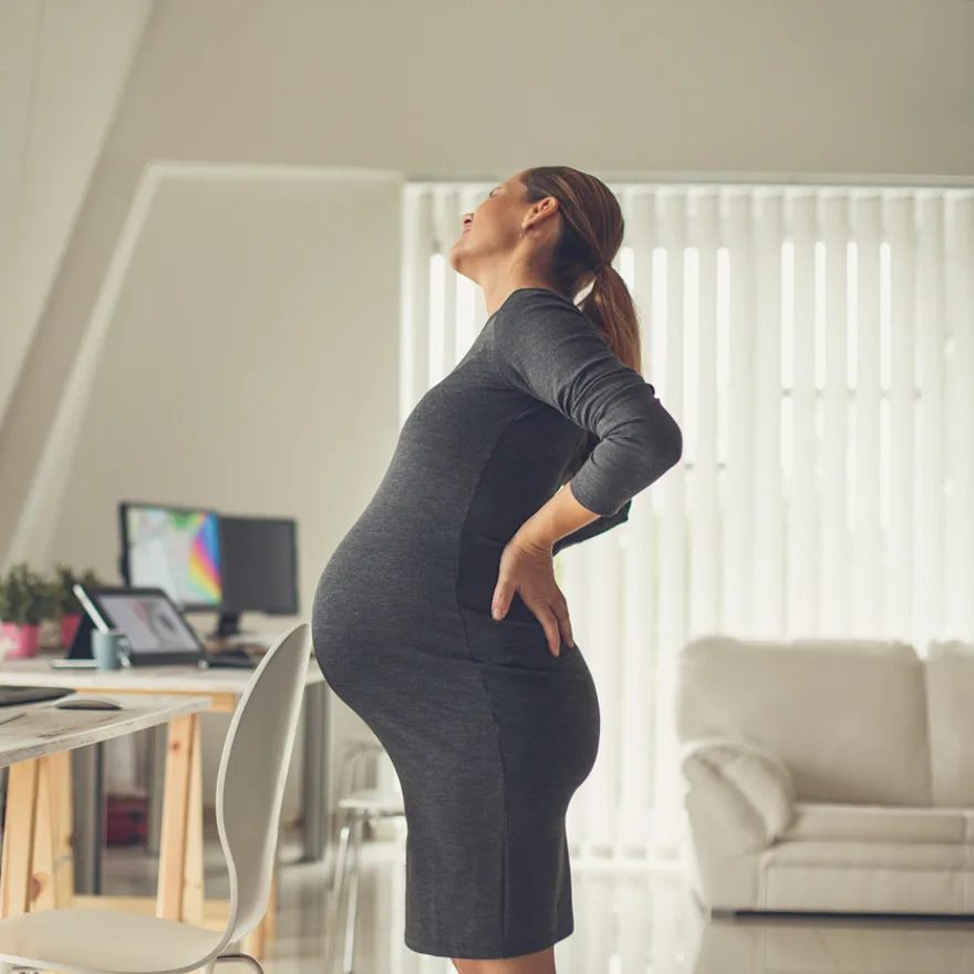 Pregnant woman experiencing SI joint pain while working at standing desk in bright modern office space