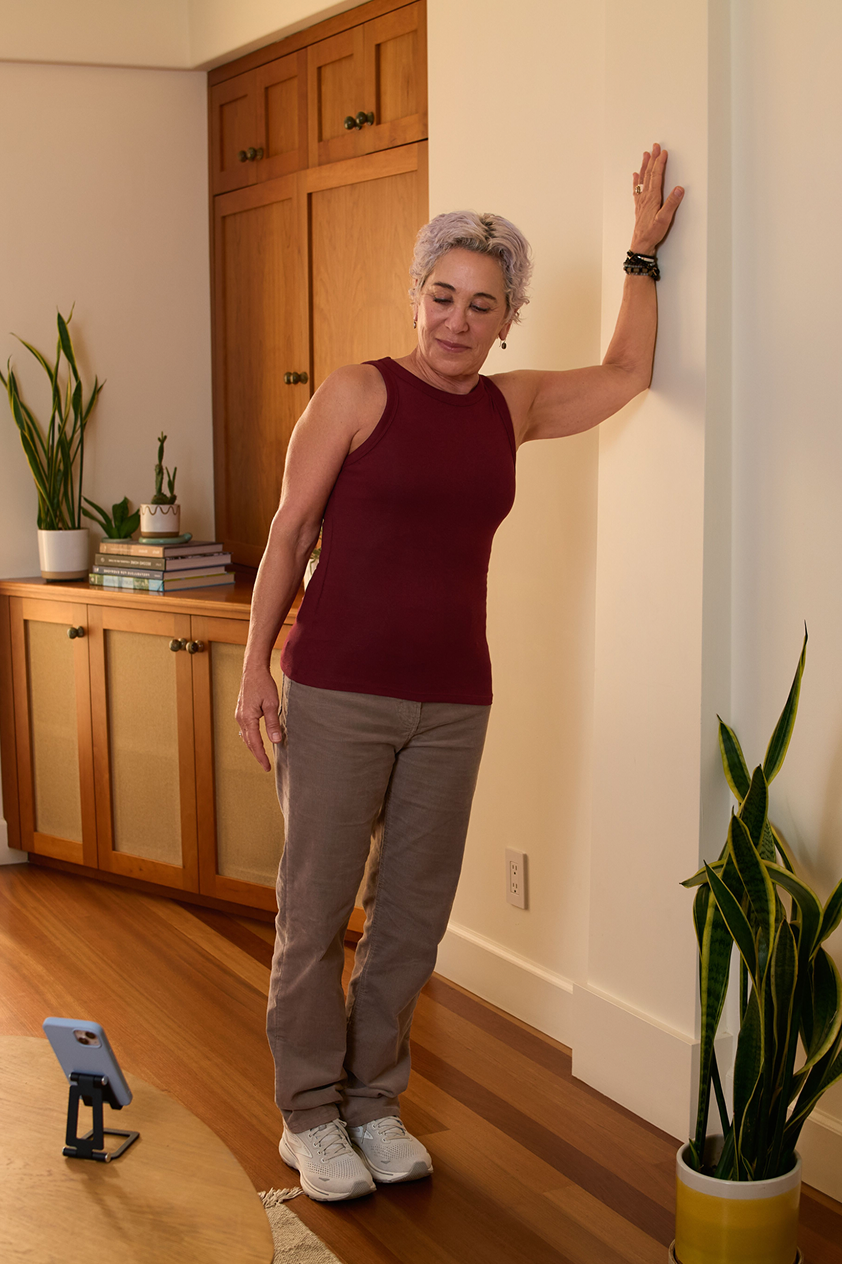 An older woman does a strength exercise with her forearm resting against a wall. Her phone is on a device stand nearby. A screen shows a confirmation for a video visit with her physical therapist.