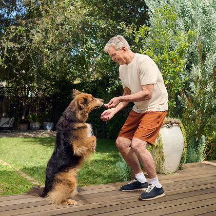 An older man is bending down to pet his dog in his sunlit backyard.