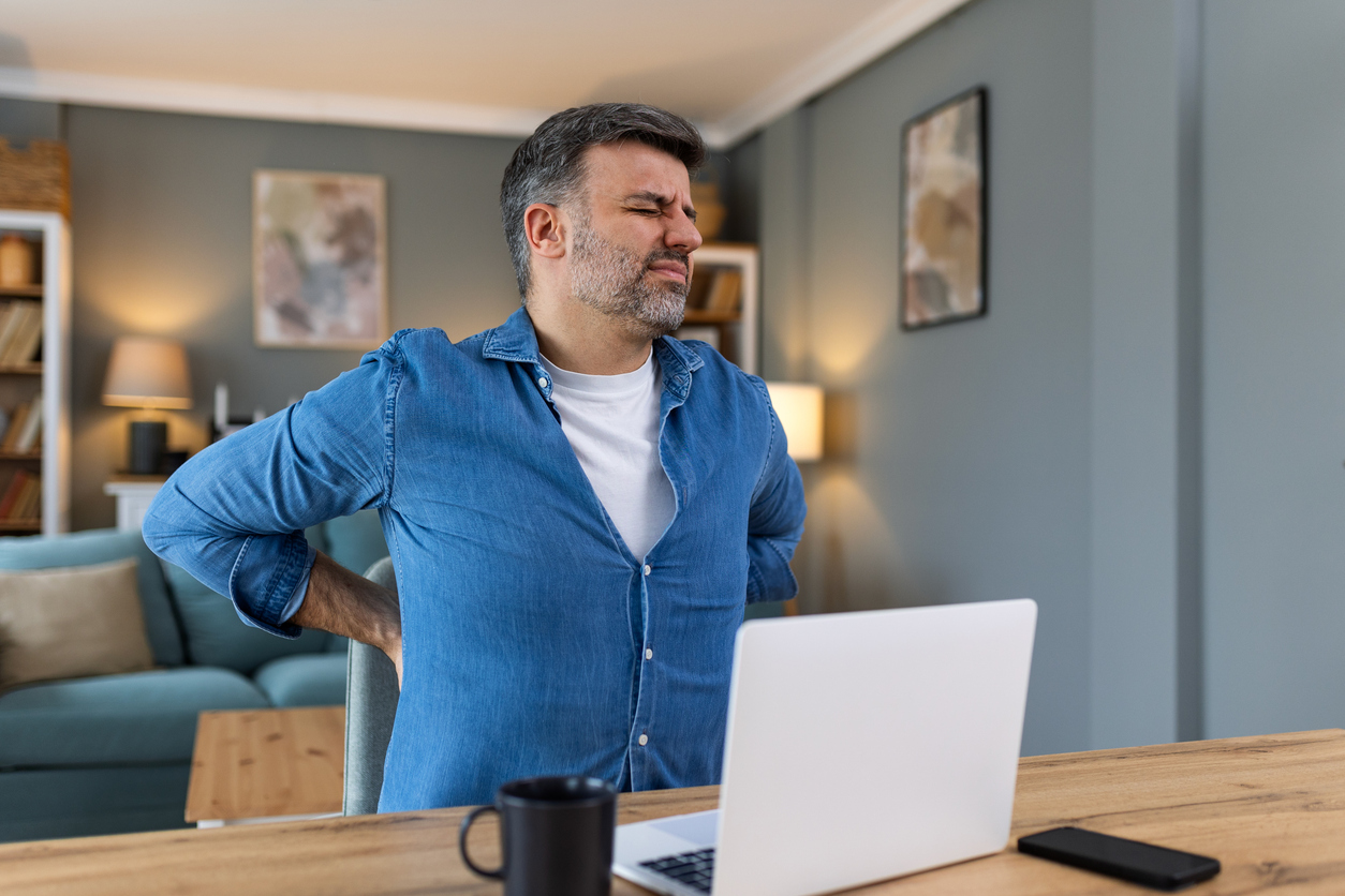 Person in blue denim shirt grimacing while holding lower back in pain while working at desk with laptop in home office setting