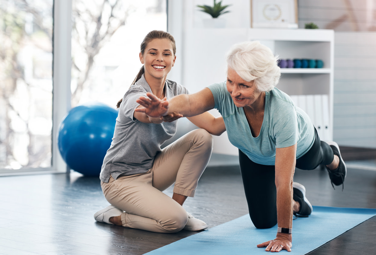 Physical therapist assists senior woman with balance exercise on yoga mat for physical therapy after back surgery.