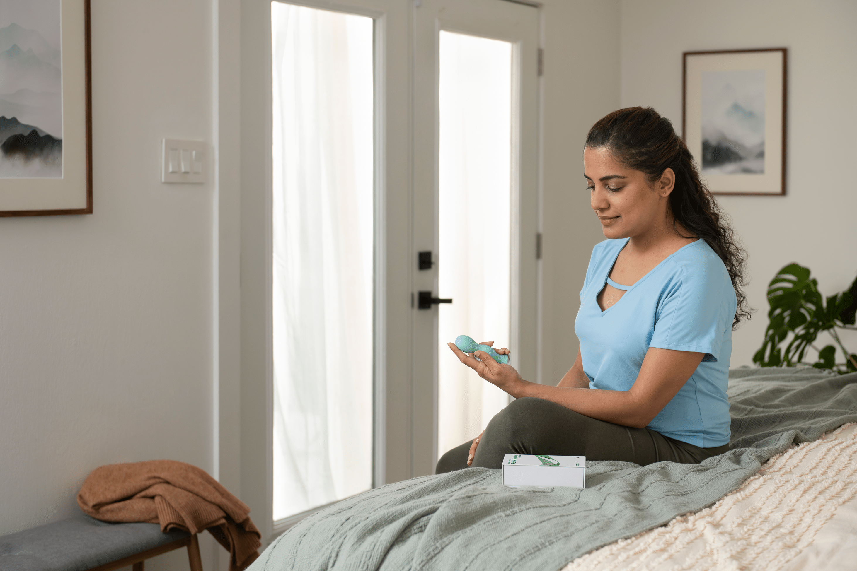 Image of a woman sitting on the edge of a bed, holding a pelvic trainer in her hand