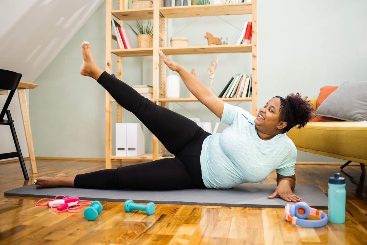 Woman doing gluteus medius exercises on exercise mat at home, surrounded by colorful fitness equipment and wooden shelving