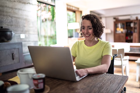 A woman in a wheelchair using a laptop at a table. A woman in a wheelchair using a laptop at a table.