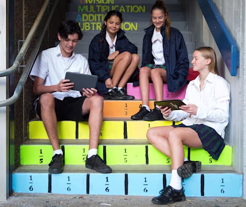 Four students sitting on colourful steps in a school setting looking at their tablets and talking Four students sitting on colourful steps in a school setting looking at their tablets and talking