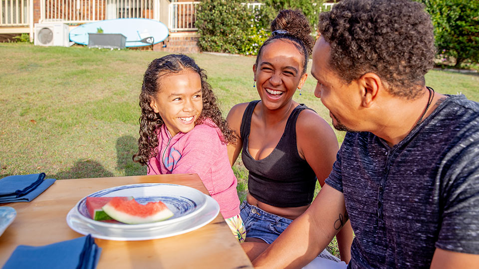 A young family laugh together in the backyard with the remains of a watermelon slice sitting on a plate in front of them