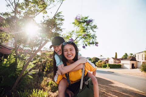 Teenage girls hanging out together walking along a neighbourhood street. One girl is giving the other a piggyback as they laugh together enjoying summer. Teenage girls hanging out together walking along a neighbourhood street. One girl is giving the other a piggyback as they laugh together enjoying summer.