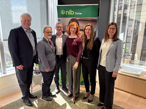 Six people from nib, People With Disability Australia and Down Syndrome Australia stand in an office space in front of a "nibThrive" banner. City buildings and indoor plants are visible in the background. Six people from nib, People With Disability Australia and Down Syndrome Australia stand in an office space in front of a "nibThrive" banner. City buildings and indoor plants are visible in the background.