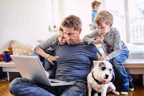 A man sits on the ground looking at his laptop with his two sons and a dog surrounding him A man sits on the ground looking at his laptop with his two sons and a dog surrounding him