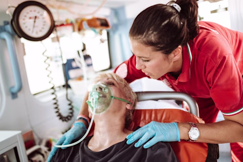 A woman sits in an ambulance with an oxygen mask on, while a paramedic holds her shoulders