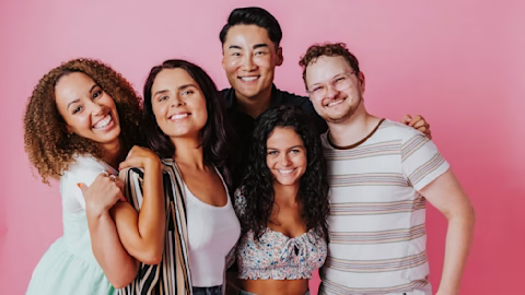 A group of young adults smiling in front of a pink backdrop A group of young adults smiling in front of a pink backdrop