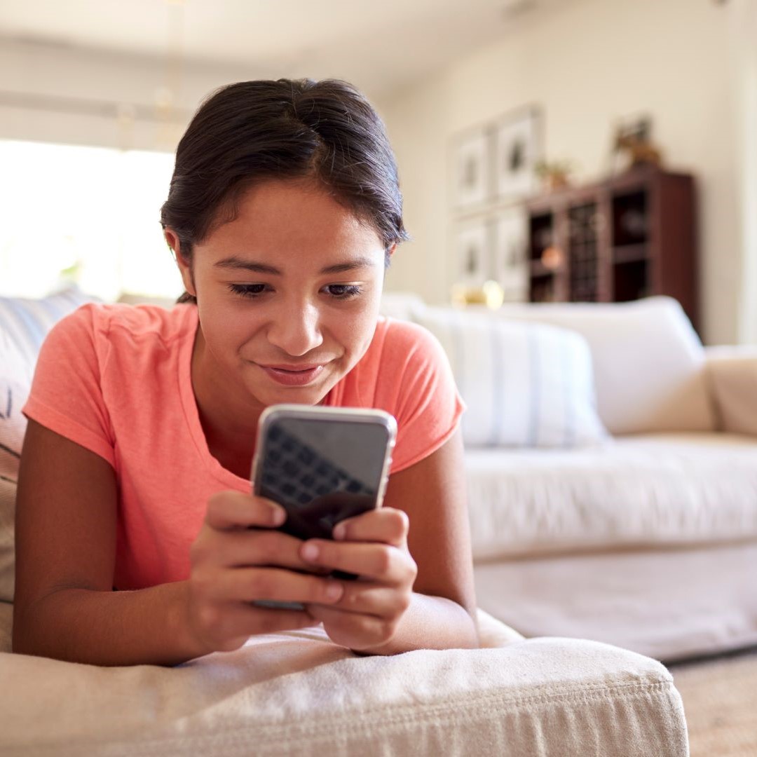 Young teen girl in peach top on couch smiling at phone