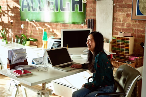 A person of Asian heritage sitting at a desk, using a laptop, laughing. A person of Asian heritage sitting at a desk, using a laptop, laughing.