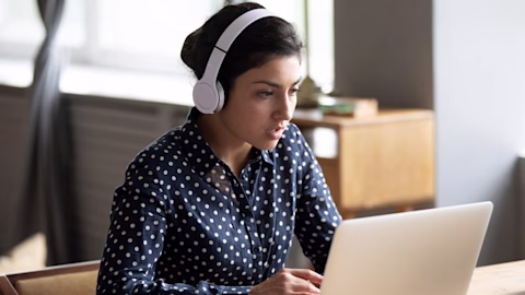 Woman wearing headphones sitting at her desk working on a laptop Woman wearing headphones sitting at her desk working on a laptop