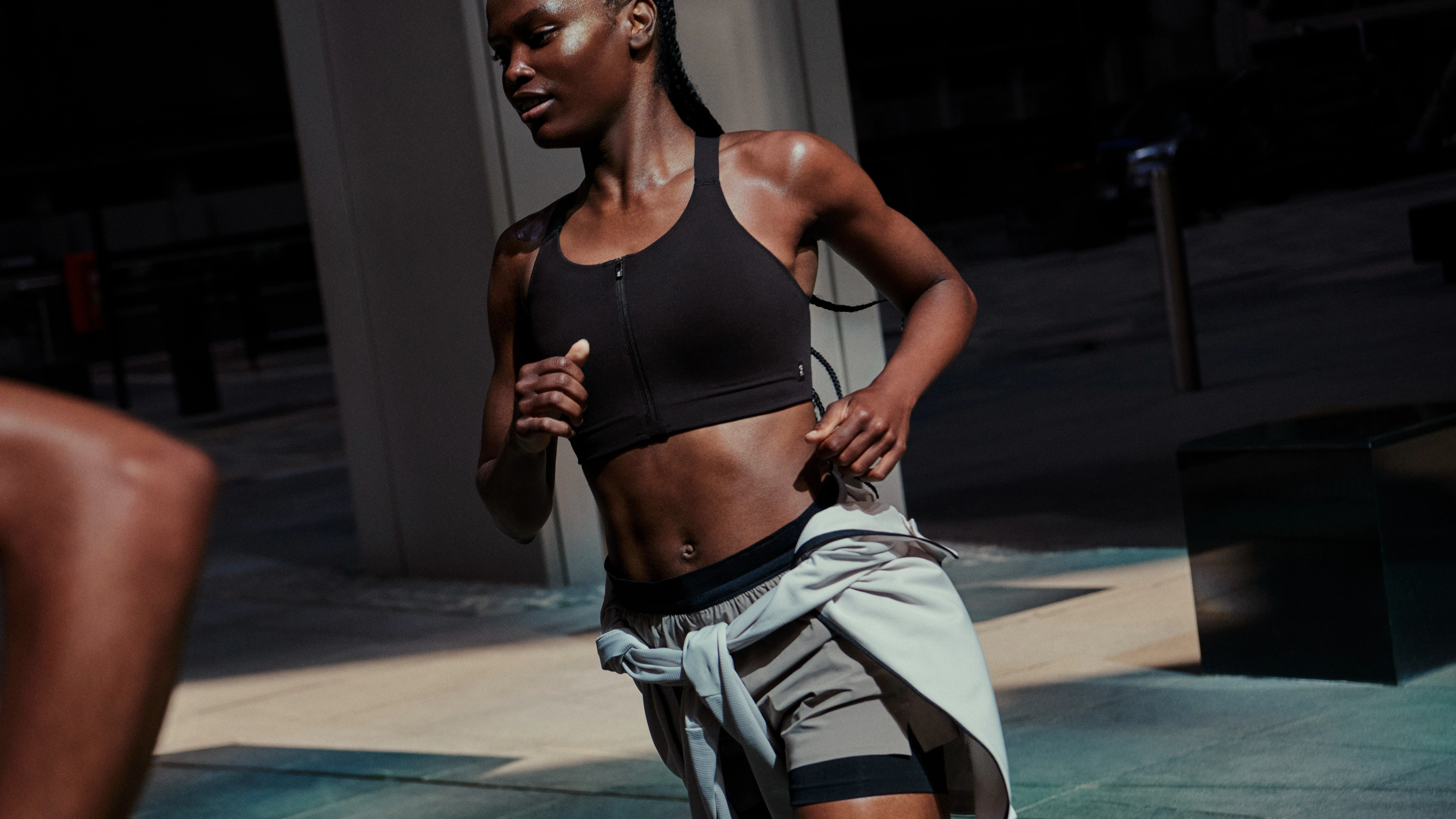 A woman running outside, wearing an On sports bra and shorts with a sweatshirt tied around her waist.