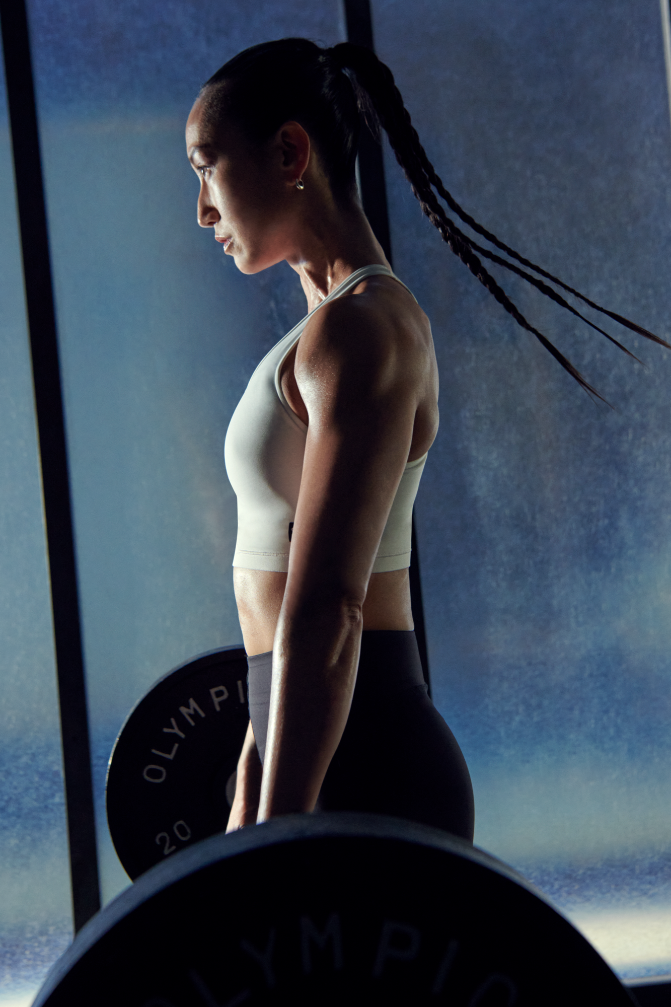Side view of a woman lifting a barbell in a gym, wearing a white sports bra and black leggings.