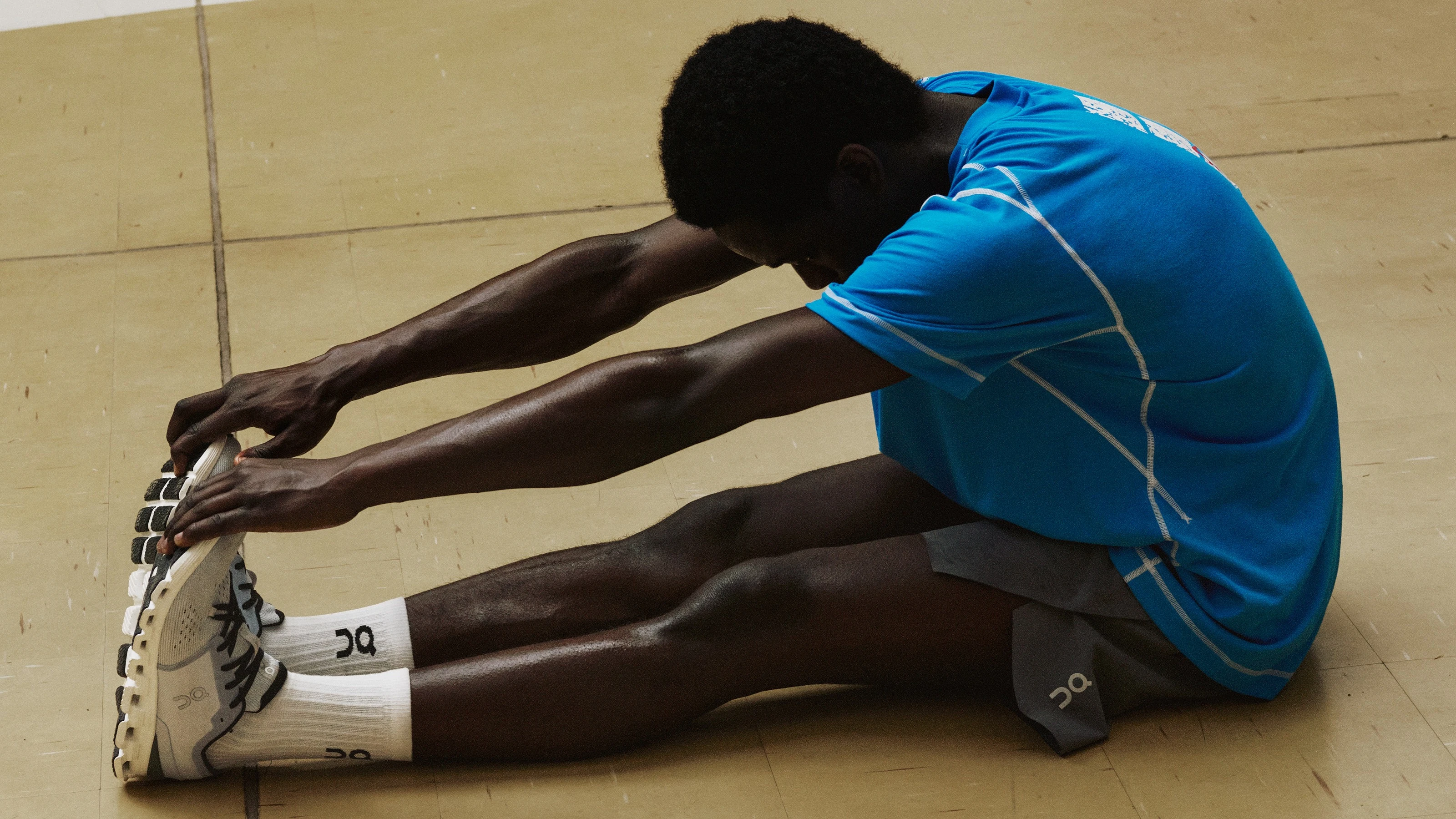 A man wearing On athletic gear while performing a seated hamstring stretch in an indoor court.