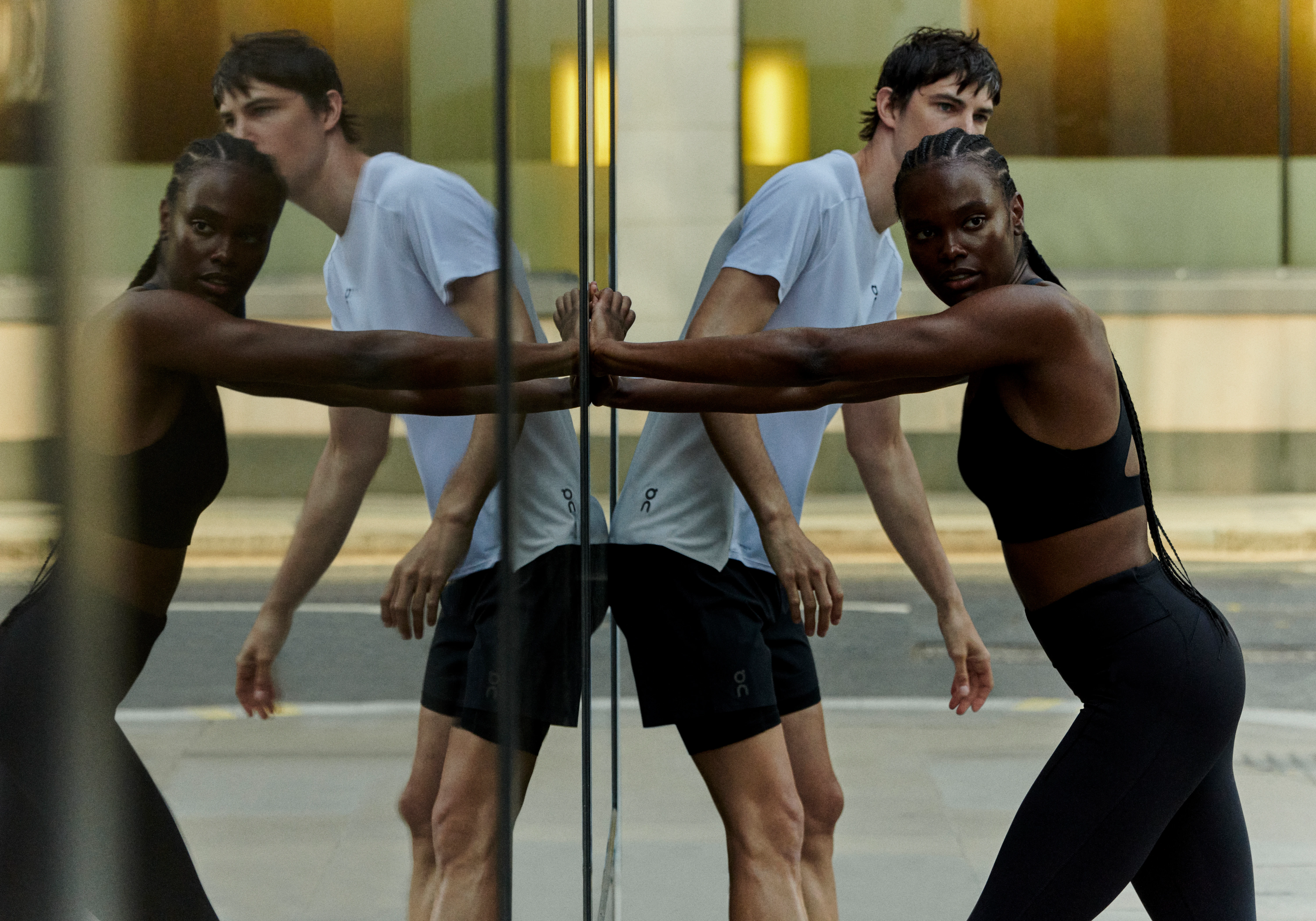 Two athletes wearing On performance gear stretch outside while leaning against a reflective surface.