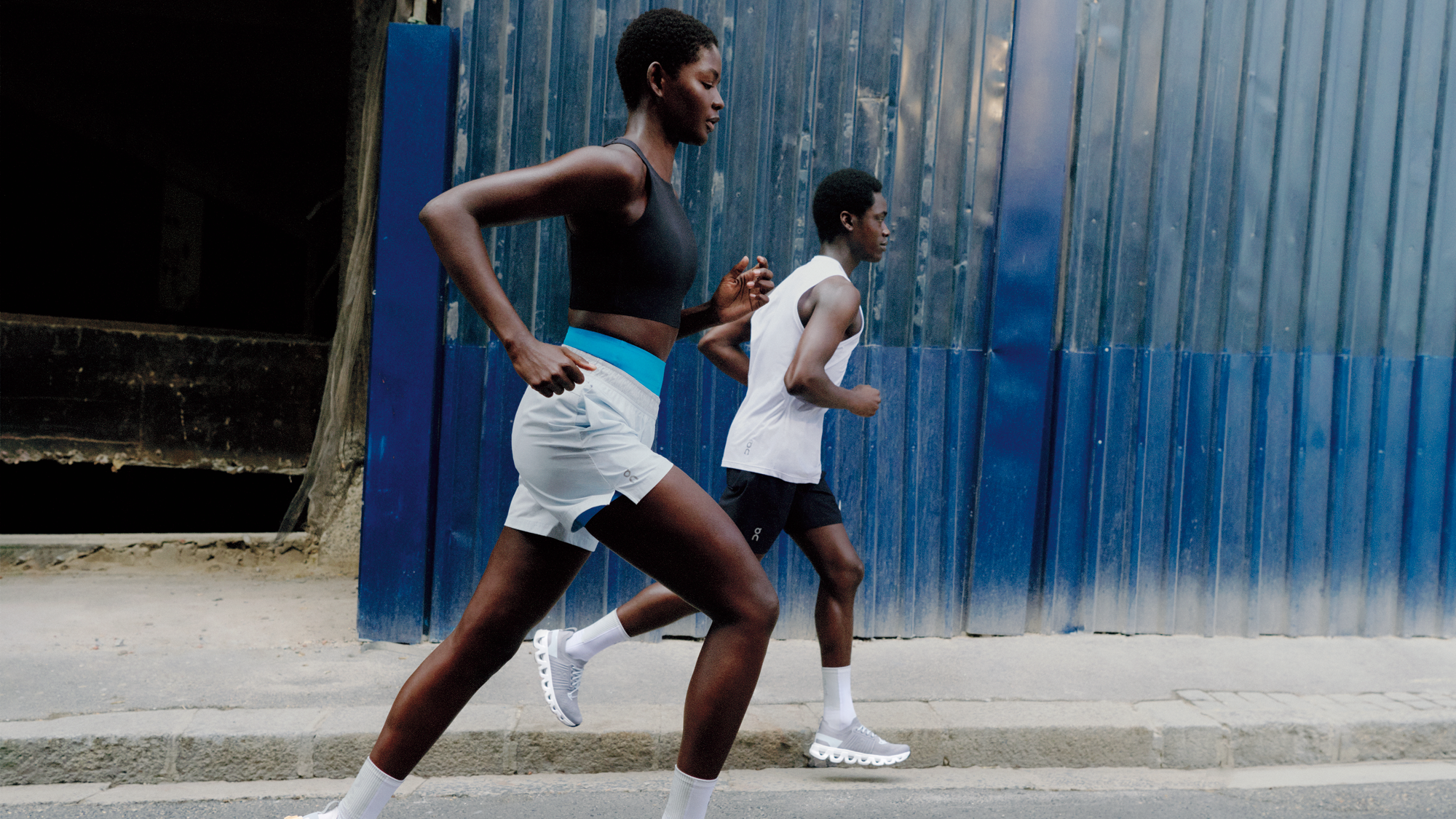 Dos personas con equipación de running corren al aire libre. La mujer más cerca de la cámara lleva pantalones cortos blancos y un sujetador deportivo negro. 