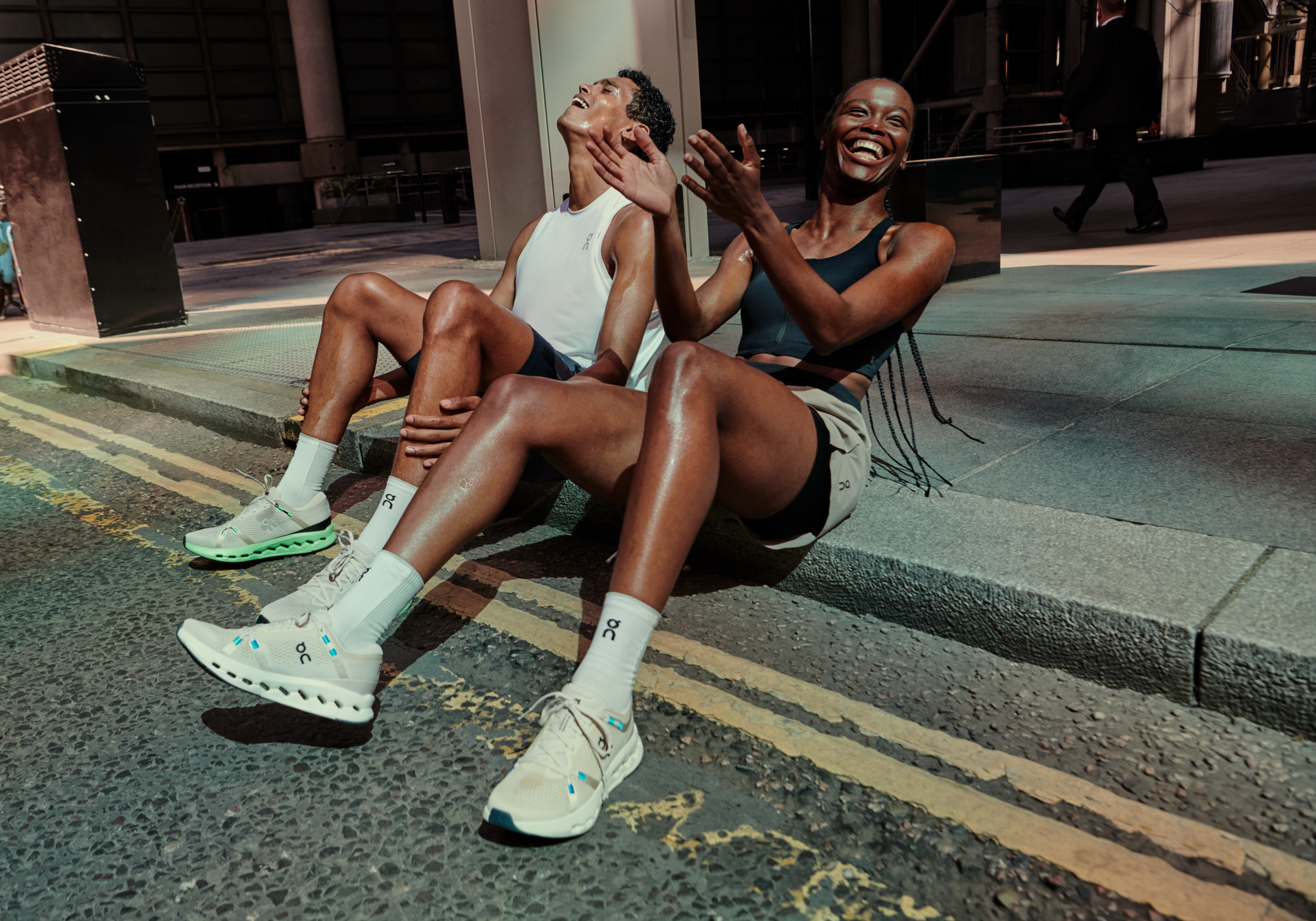 Two athletes sit side by side on the edge of a city sidewalk after their workout, wearing On athletic shoes, socks, and performance apparel.