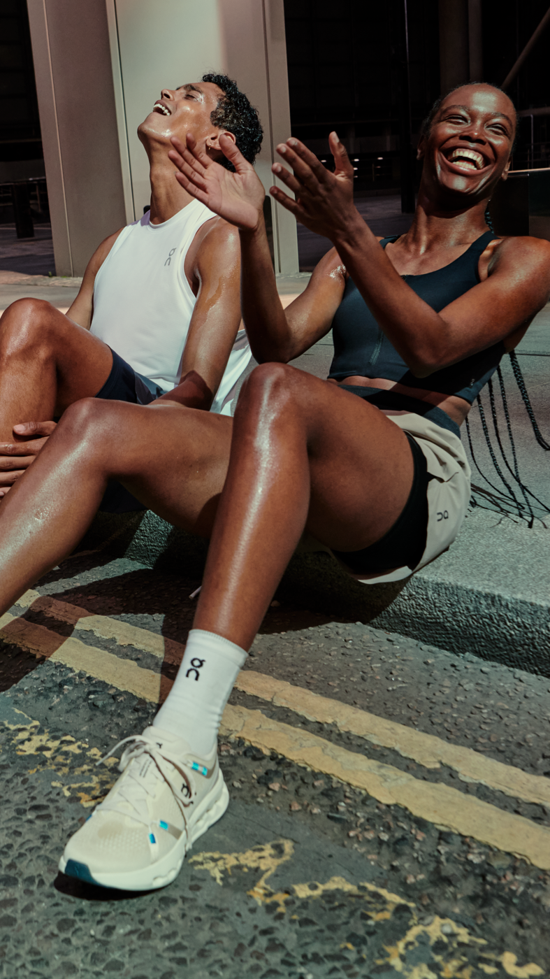 Two athletes sit side by side on the edge of a city sidewalk after their workout, wearing On athletic shoes, socks, and performance apparel.