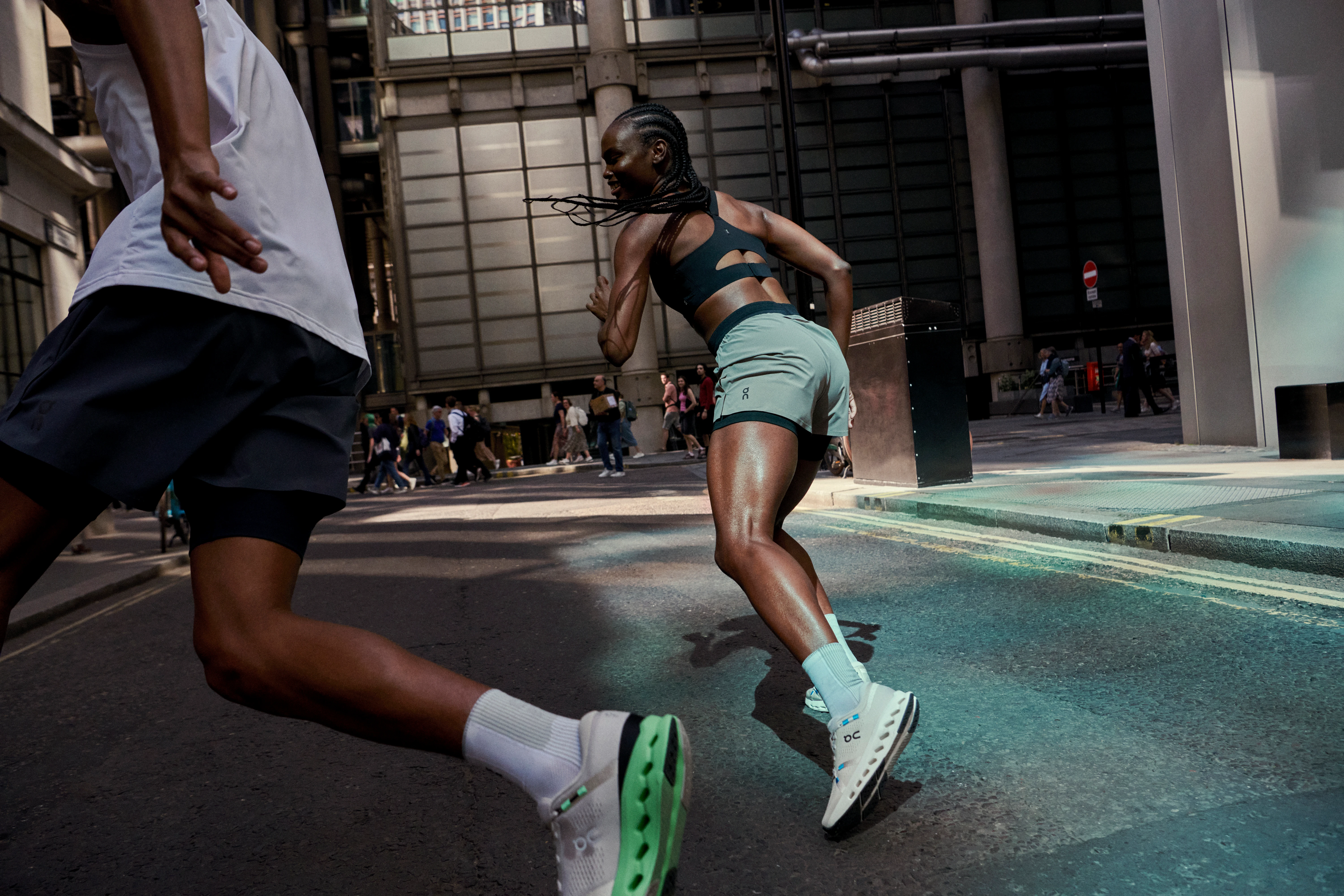 Two runners running through a city street, wearing athletic gear and On running shoes.