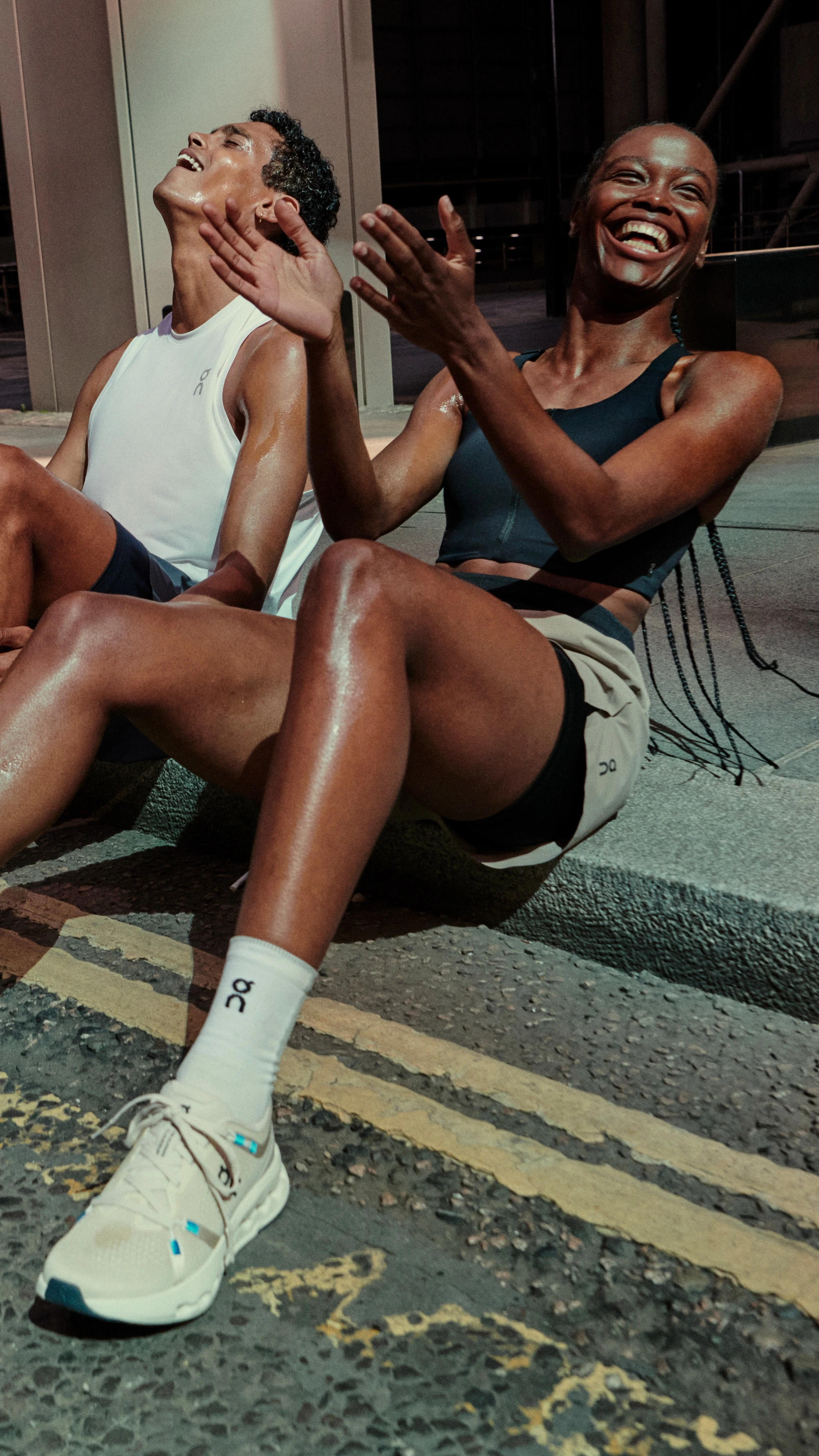A man and a woman in On apparel sitting on the curb laughing.