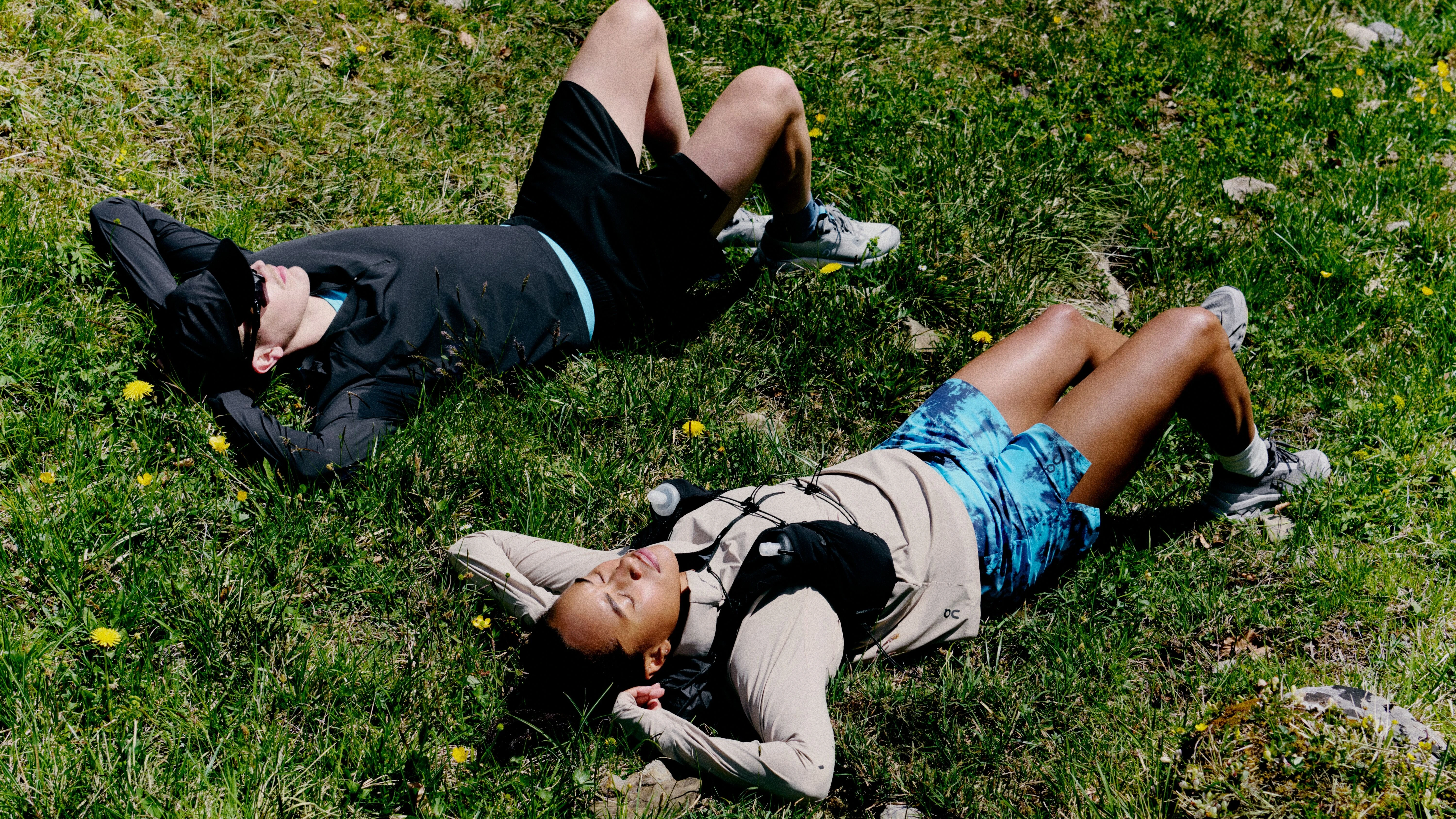Hikers resting on a grassy hill with hands overhead and eyes closed.