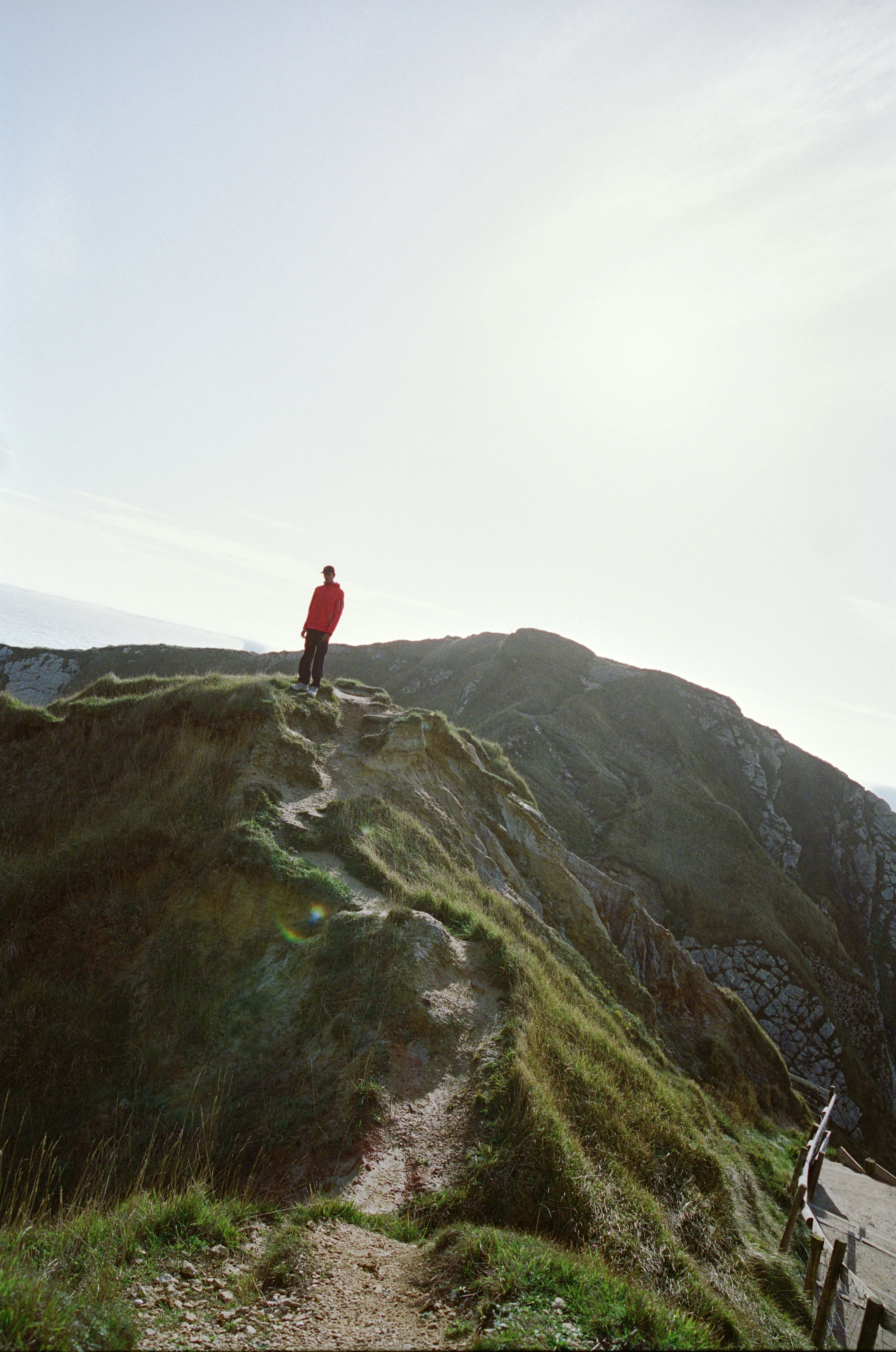 A solo hiker in a red jacket pictured at a distance along a grassy ridgeline.