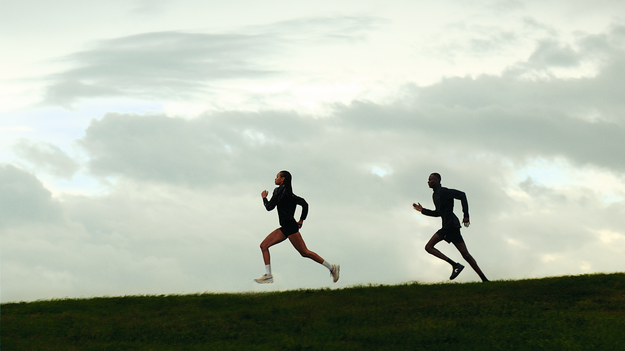 Two runners in athletic gear sprinting uphill against a cloudy sky.
